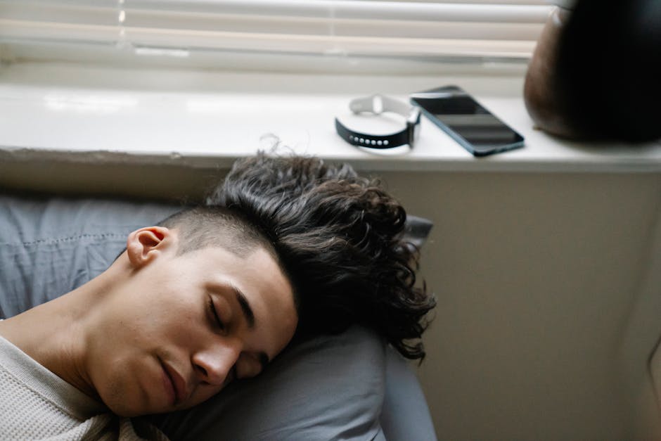A young man with curly hair sleeping in a modern bedroom with a smartphone and wristwatch visible.