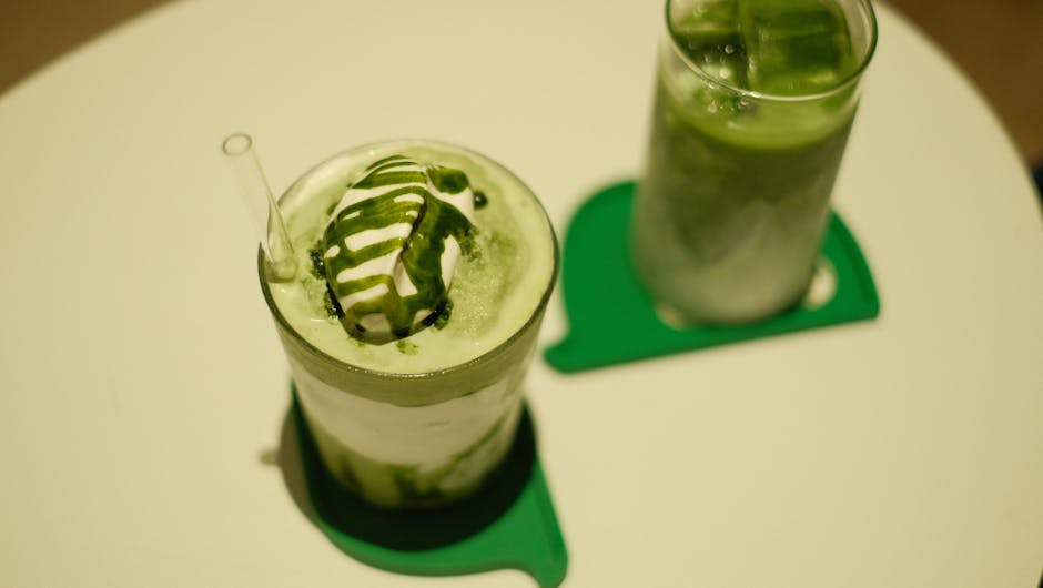Two iced matcha lattes topped with cream and matcha drizzle on a table.