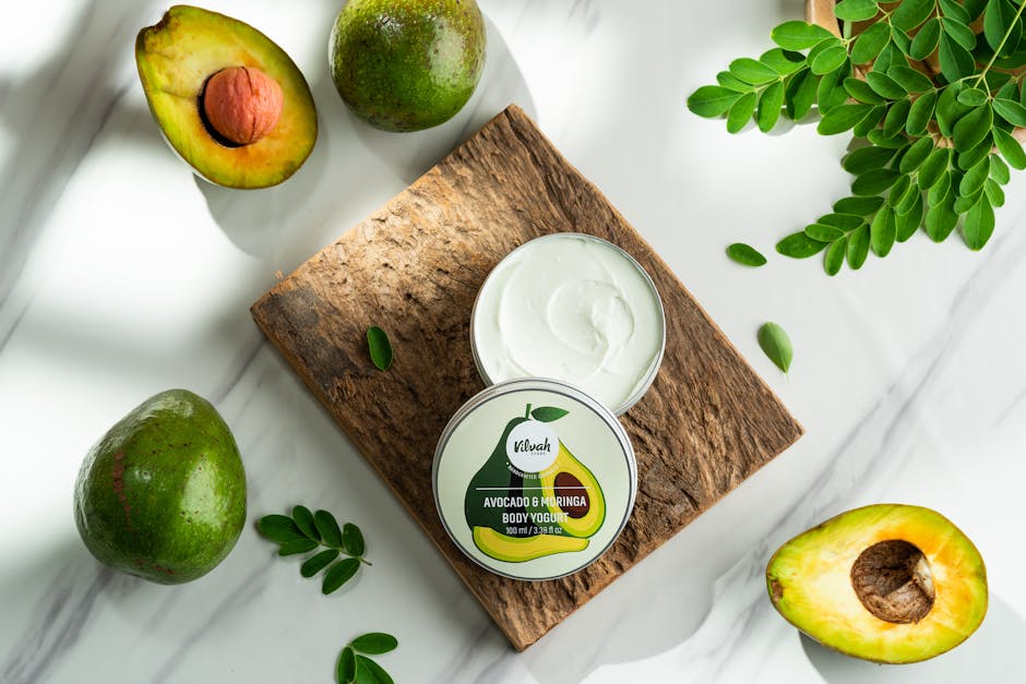 Top view of avocado and moringa body yogurt container on rustic wooden board surrounded by avocados and moringa leaves.
