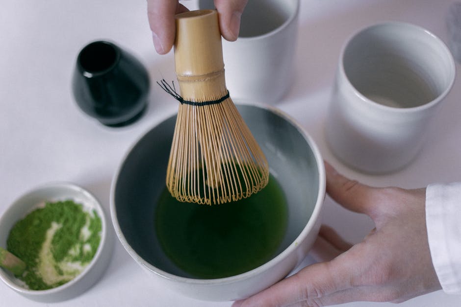 Close-up of hands using a bamboo whisk to prepare matcha tea in a ceramic bowl.