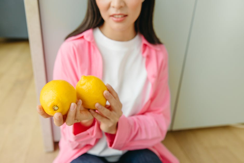 A woman in a pink shirt holds two fresh lemons indoors, symbolizing health and nutrition.