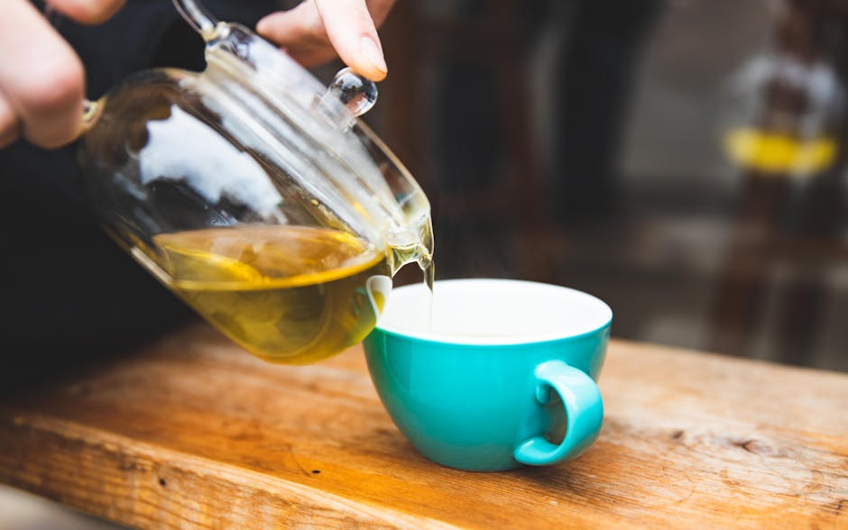 A hand pours green tea from a glass teapot into a turquoise cup placed on a rustic wooden table.