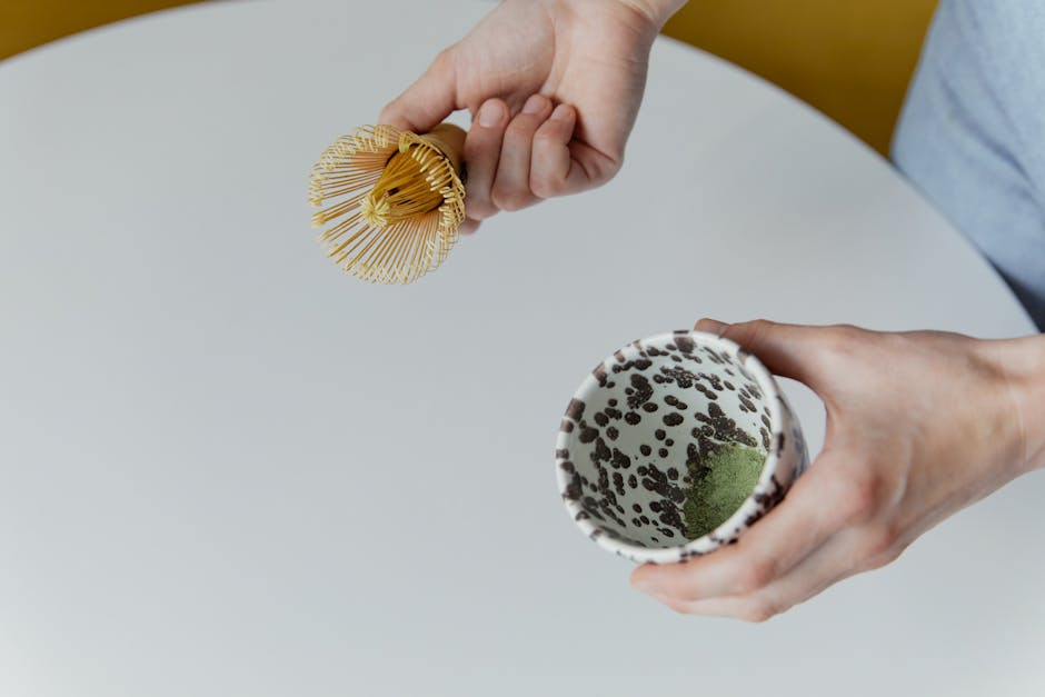Person preparing matcha tea in a patterned mug with a bamboo whisk on a white surface.