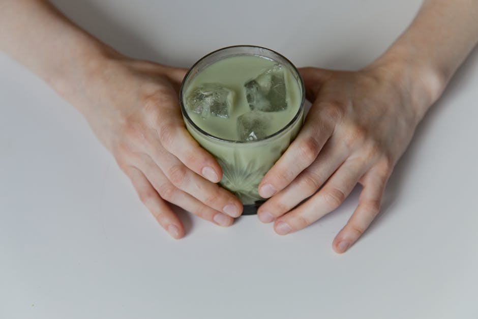 Close-up of a hand holding a glass of iced matcha latte on a white surface.