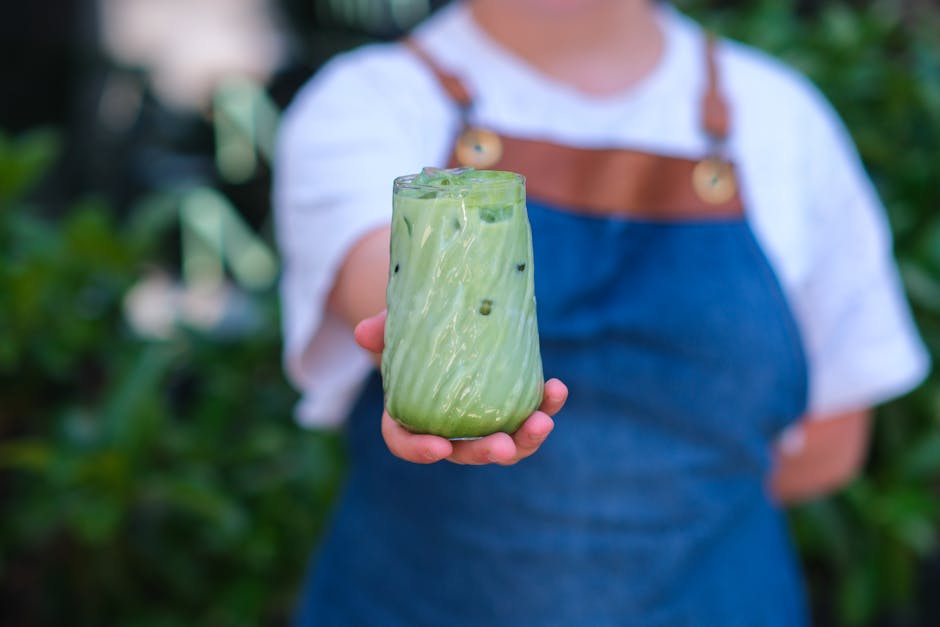 A barista holds out a glass of iced matcha latte with a blurred garden background.