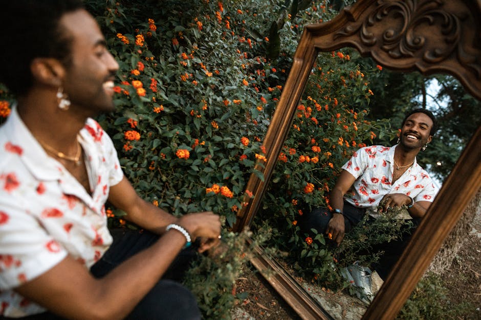 Smiling man sitting outdoors beside a mirror, reflecting in a floral garden.
