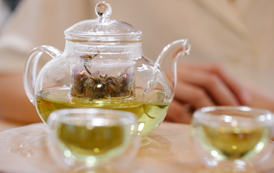 A close-up of a glass teapot and cups filled with herbal green tea on a wooden table.
